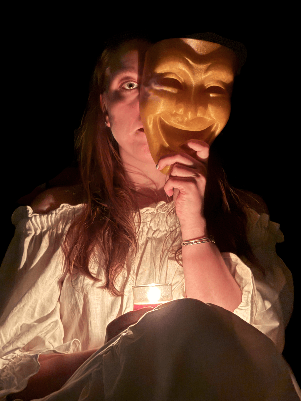 A candlelit photo of -k. hiding half of her face behind a golden comedy mask.